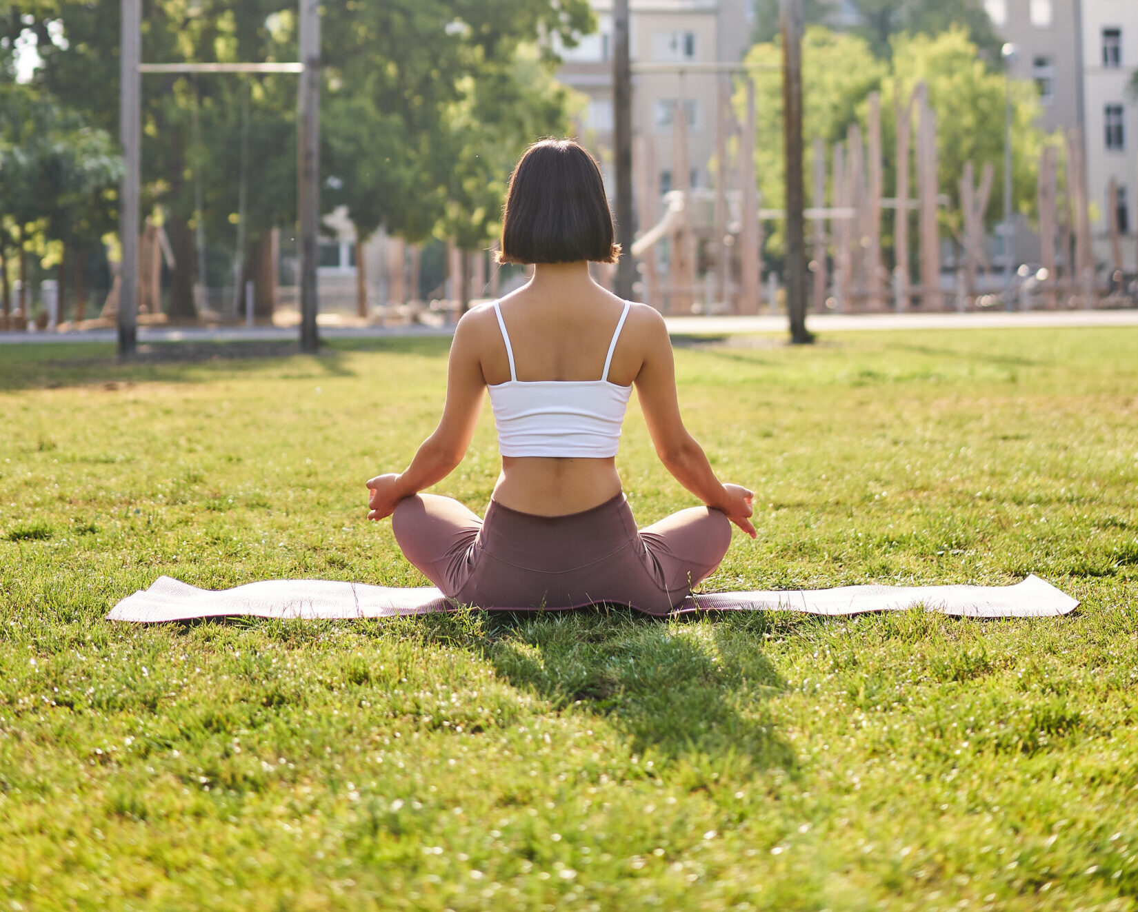 Rear view of young woman meditating in morning park, relaxing, breathing practice and wellbeing training session